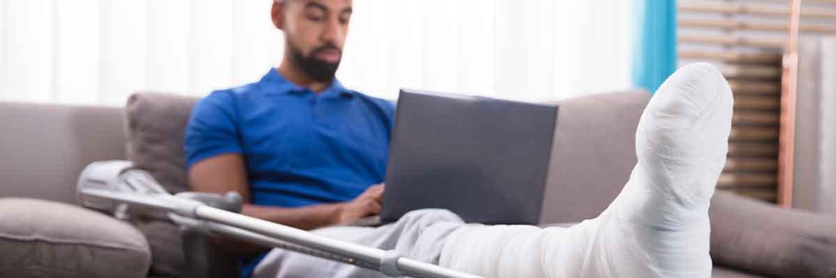 Man with a leg in a cast, sitting on a couch with a laptop in his lap.