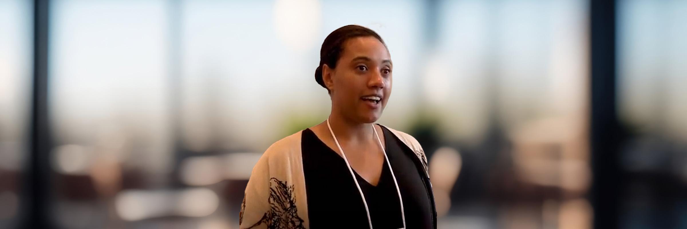 Photo of a community worker, Leah, standing indoors with a blurry background of tall window panes. 