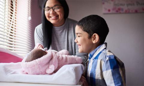 Tara stands over her baby daughter on the changing table while her son looks on. Both are smiling.