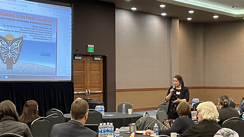 Vice Chair Loni Greninger speaking to attendees. There is a large screen on the left with a American Indian artwork.