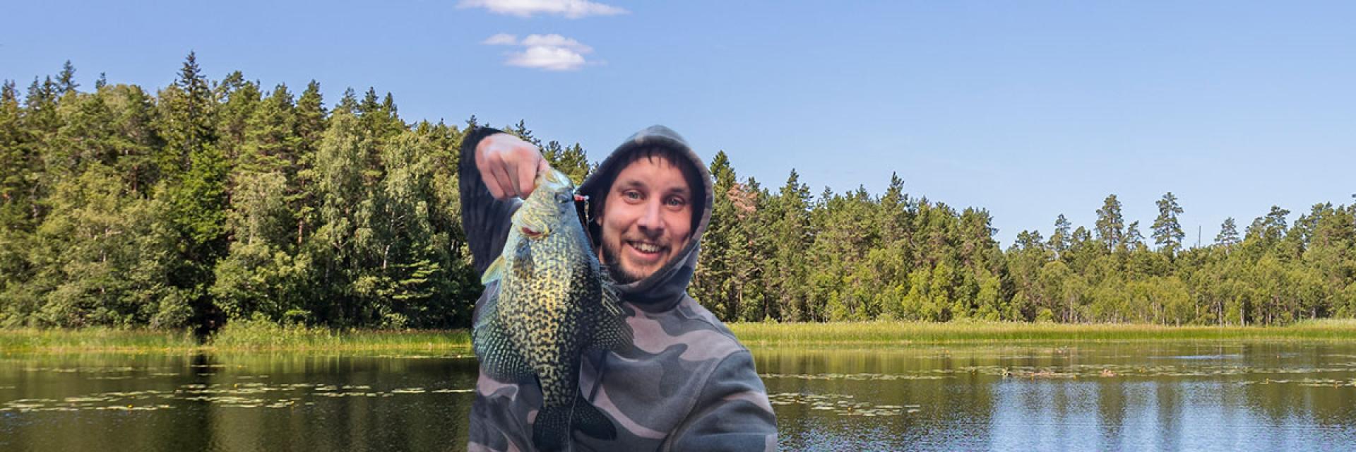 Gregory standing in front of a lake with trees across the water, holding a large fish and smiling.