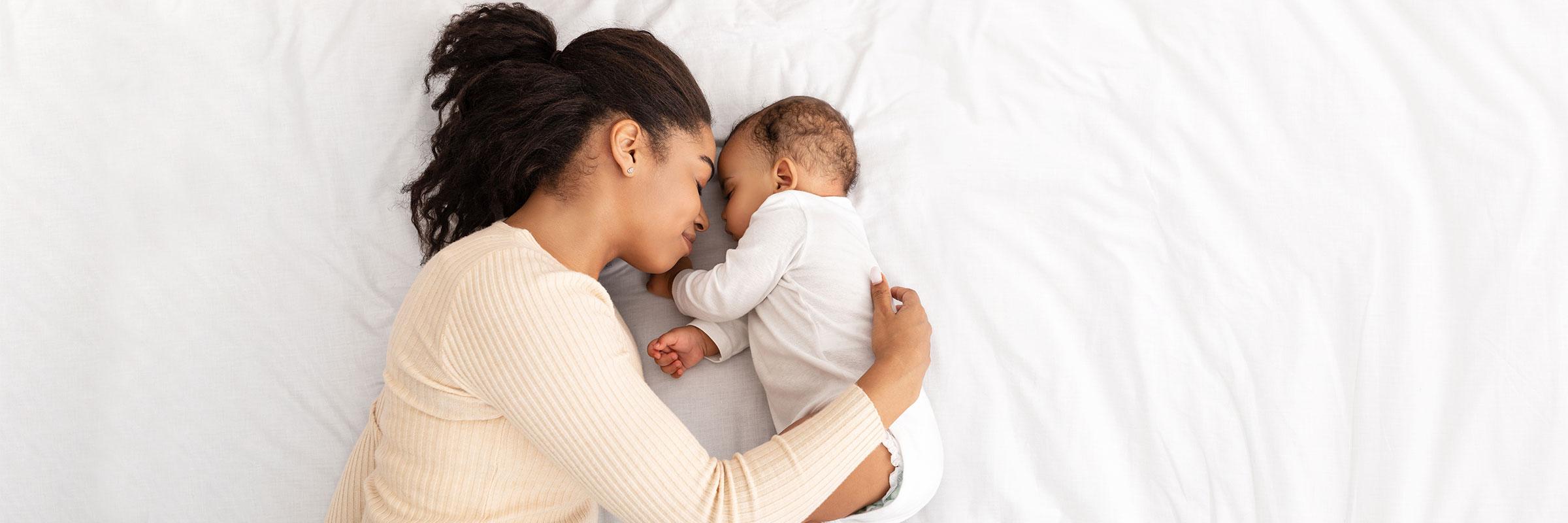 Young black mother cuddles her infant on a bed of white sheets.