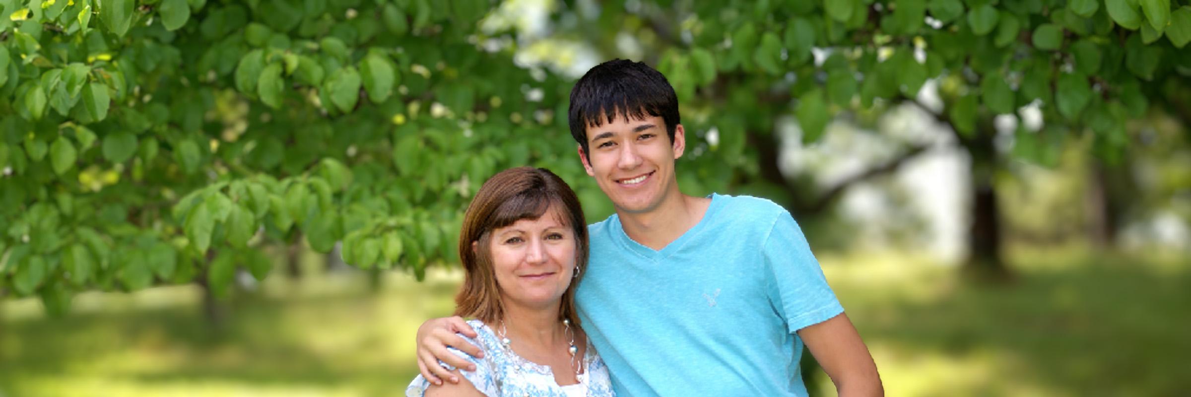 Photo of mother and son hugging each other facing camera smiling. A blurred background of outdoor scenery is behind them.