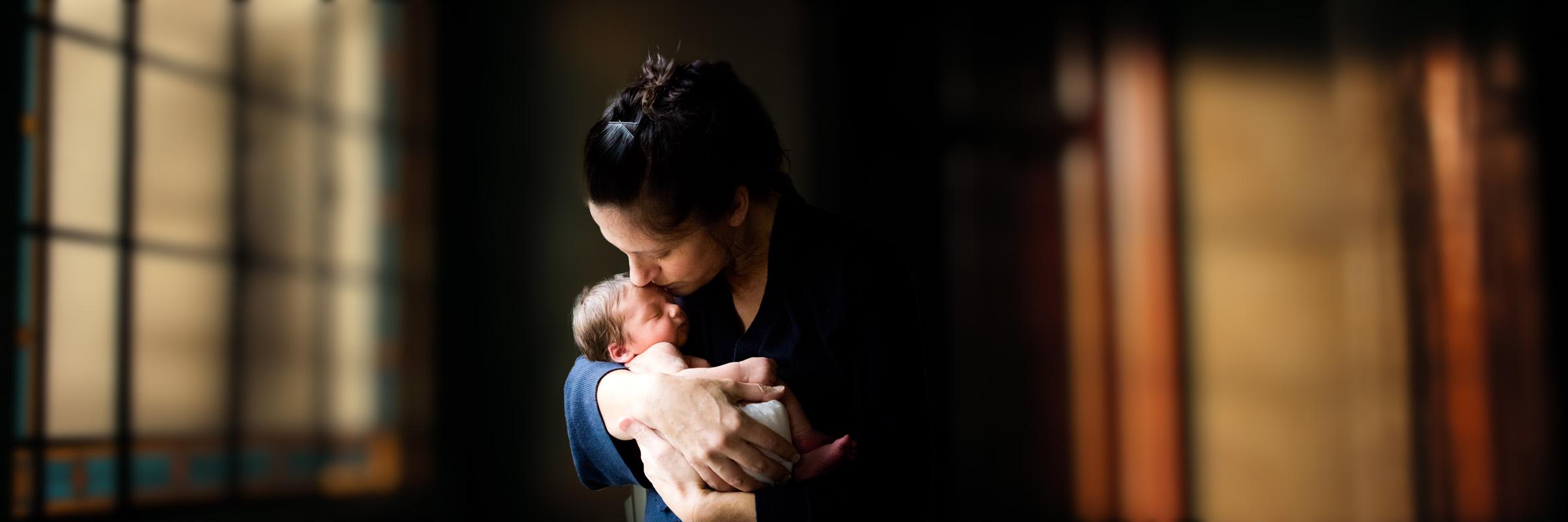 A photo of a mother holding her newborn close in both hands. They are indoors with a few closed windows behind them with daylight glowing behind.