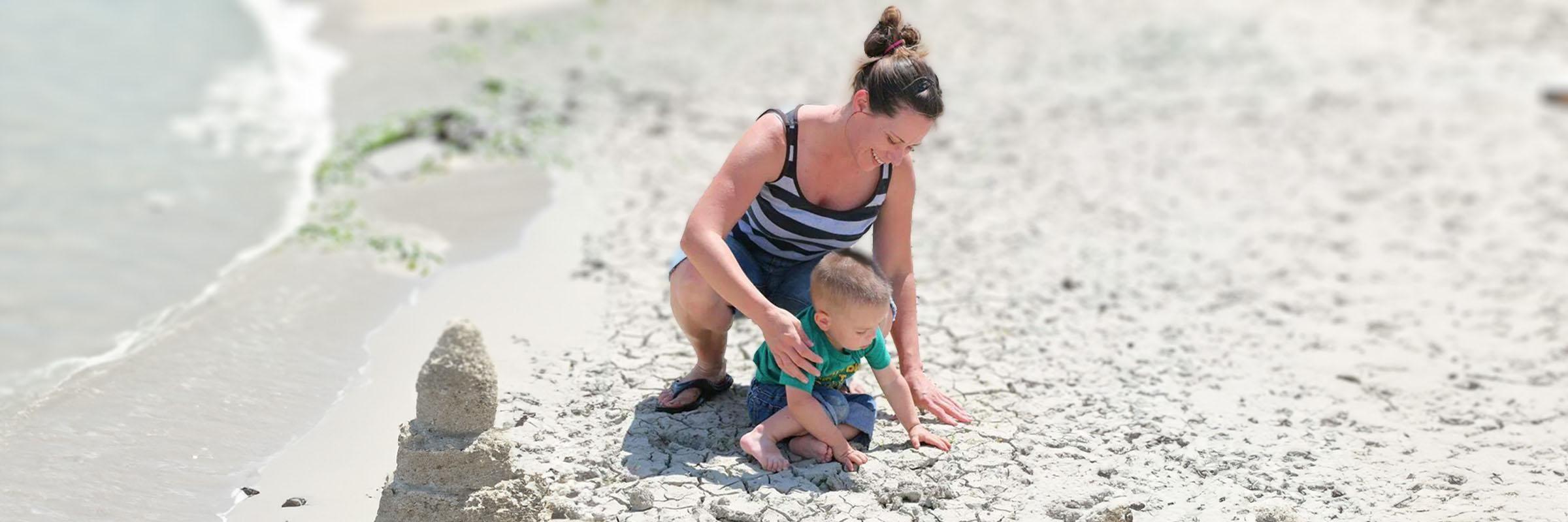 Image of an Apple Health client, Shylo, sitting in sand at the beach with the water shore nearing. Her toddler child is playing with her. 