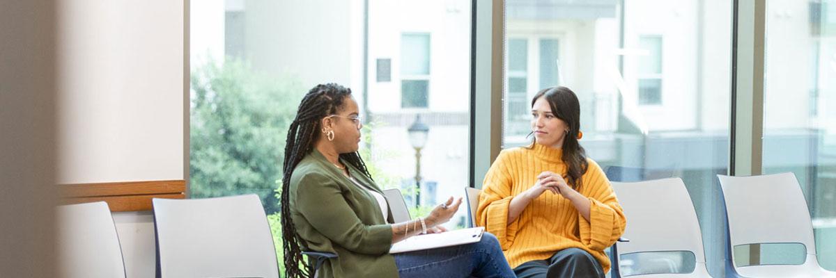 A black, woman therapist talking to a young woman in a casual setting.