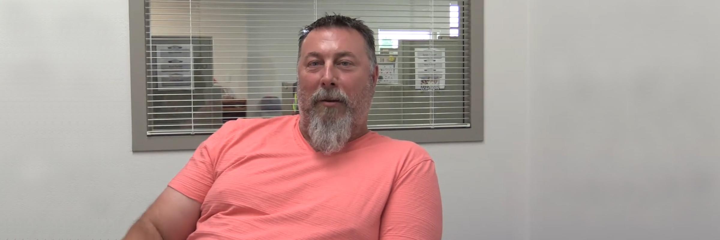 Middle aged man with salmon-colored t-shirt and a grey beard, sitting in an office with a window behind that looks into the office space.