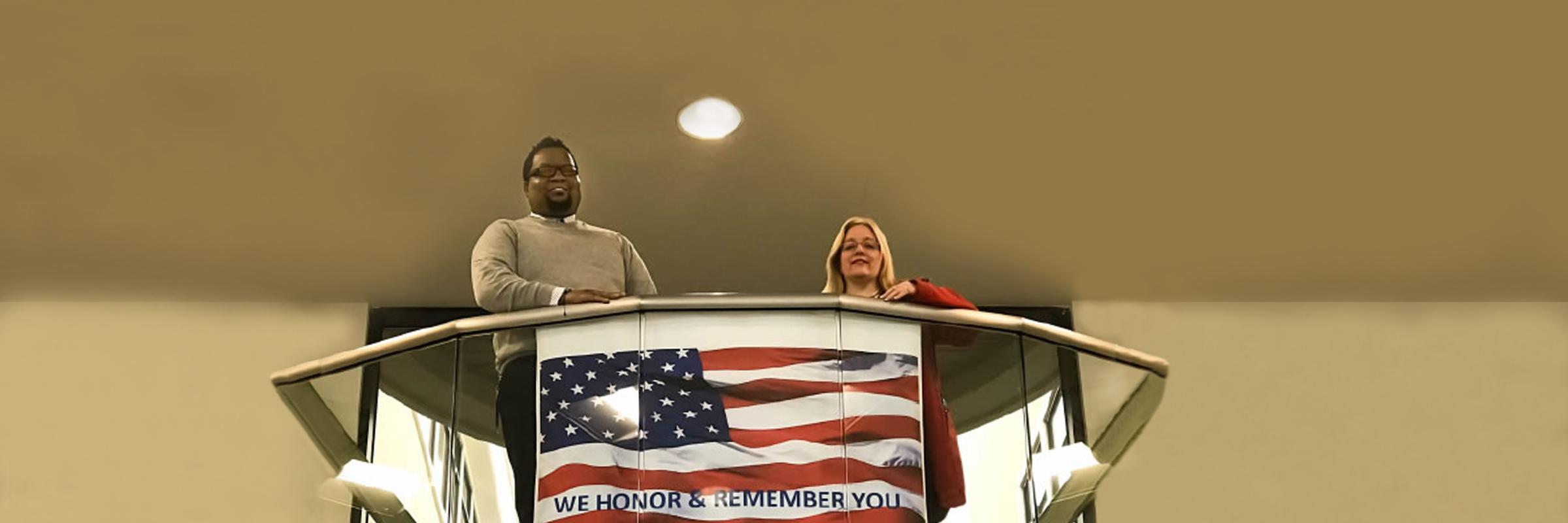 Mareco Smith and Denise Christiansen stand at a railing with a flag poster that says, "We honor and remember you."